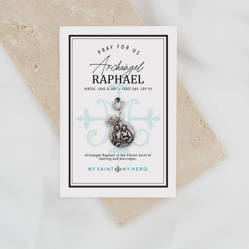 A silver-toned religious charm featuring a circular medallion with an embossed depiction of two figures, alongside a small heart-shaped charm, suspended from a lobster clasp on a light gray background.