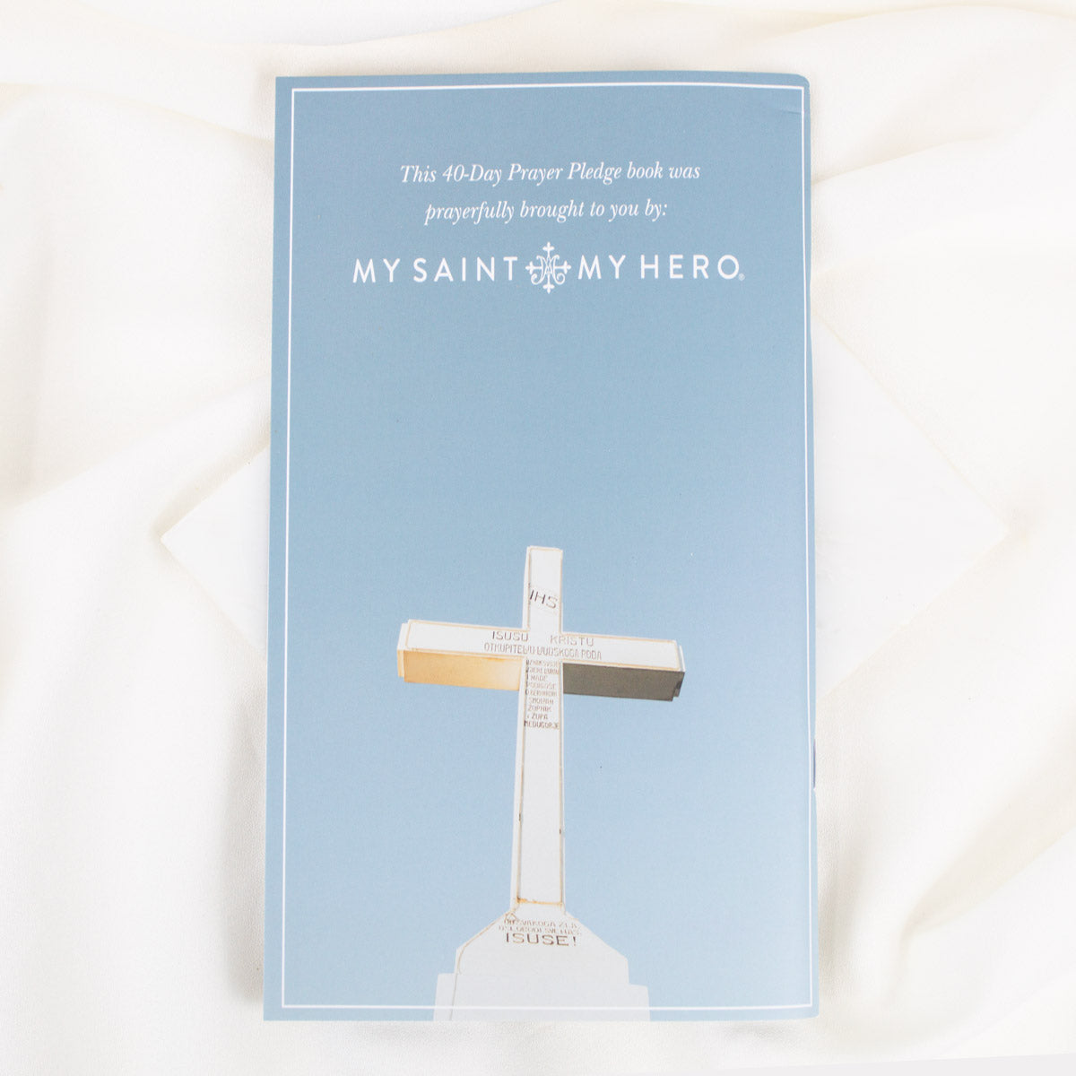 The back of the Booklet with  'MY SAINT MY HERO.' with a white cross illustration on the cover, photographed flat on a white fabric background.