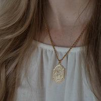 A gold pendant necklace worn by a person, featuring an oval pendant with a raised figure and decorative border, hanging from a twisted gold chain over a white top.