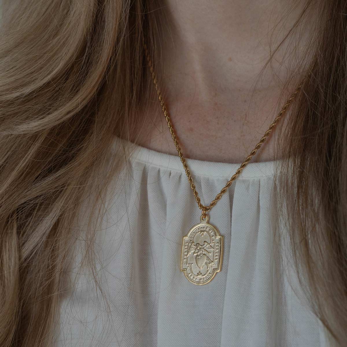 A gold pendant necklace worn by a person, featuring an oval pendant with a raised figure and decorative border, hanging from a twisted gold chain over a white top.