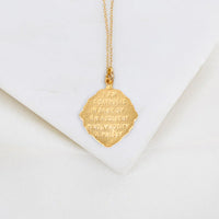 A gold pendant necklace with a textured, scalloped hexagonal charm engraved with "I am a Catholic. In case of an accident kindly notify a priest." text, displayed on a white geometric surface.