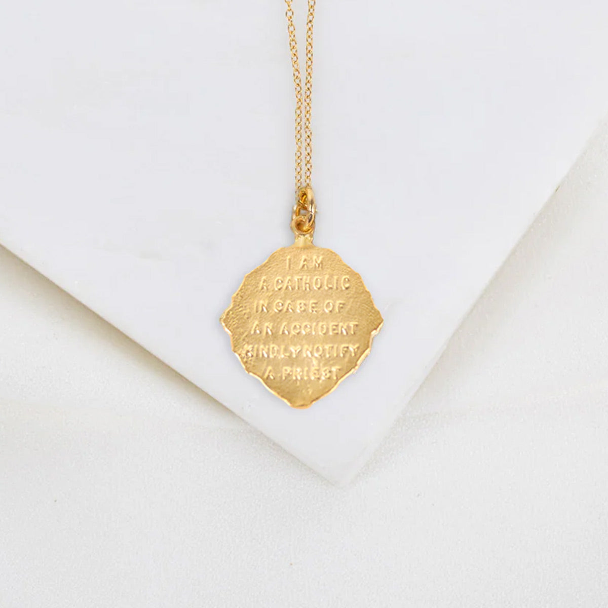 A gold pendant necklace with a textured, scalloped hexagonal charm engraved with "I am a Catholic. In case of an accident kindly notify a priest." text, displayed on a white geometric surface.