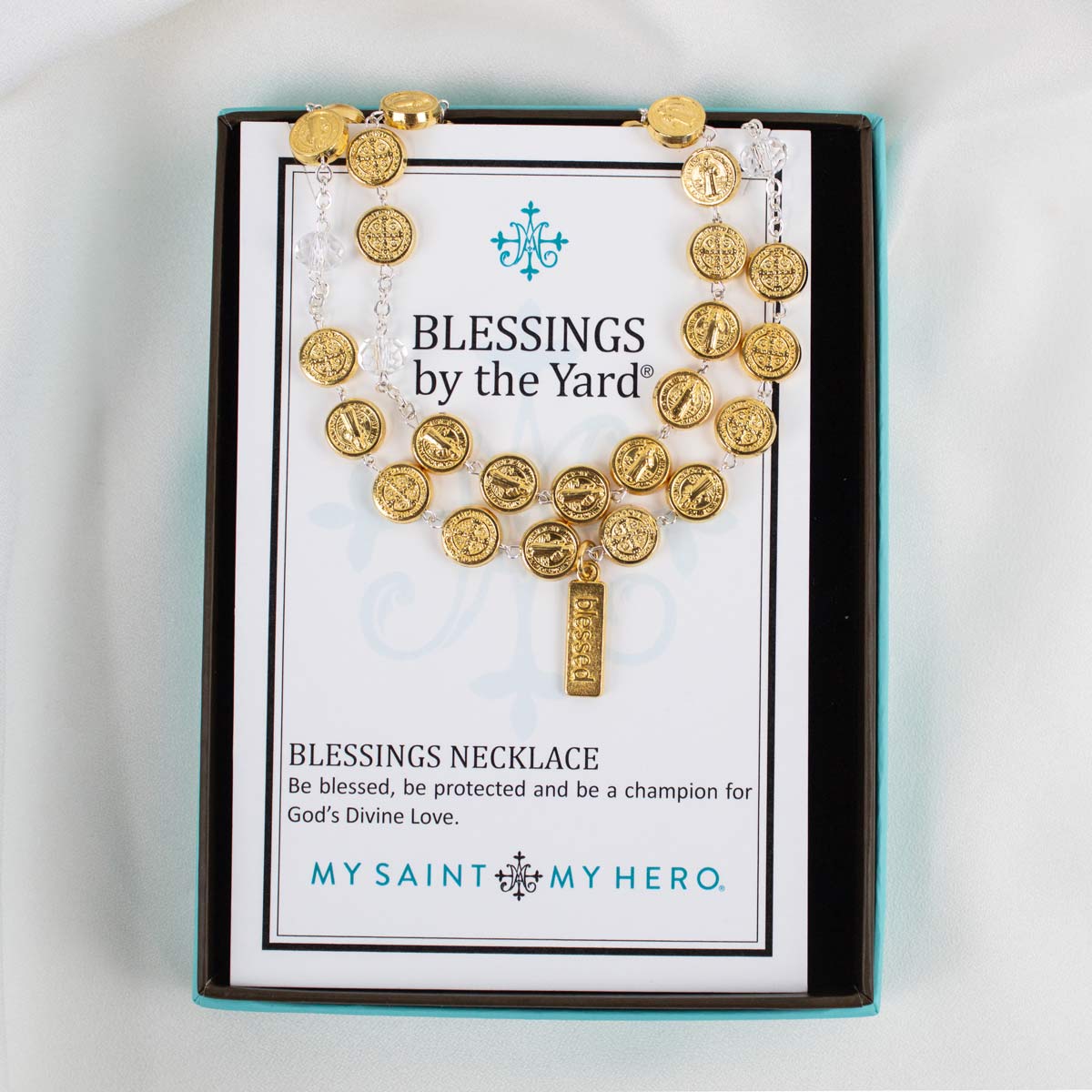 A gold Saint Benedict Blessings Necklace displayed on its branded white and black presentation card inside a turquoise box, set against a white fabric background.