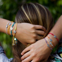 Close-up of hands wearing colorful Breathe String Benedictine Medal of Protection bracelets with a blurred green background