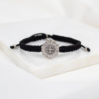 A black braided cord bracelet with a silver-toned Benedictine cross medal set with small crystals, displayed on a white surface.