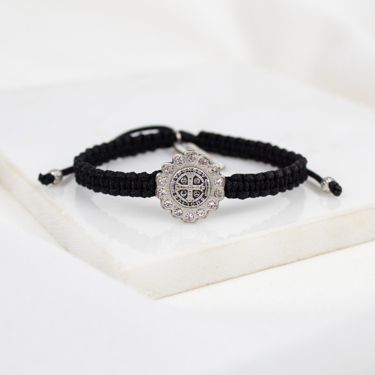 A black braided cord bracelet with a silver-toned Benedictine cross medal set with small crystals, displayed on a white surface.