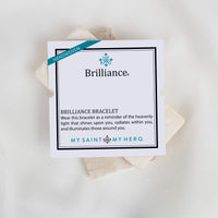 A white square card for the Brilliance bracelet, with a teal 'Handwoven' ribbon and black text, placed on top of several light beige woven fiber squares against a white background.