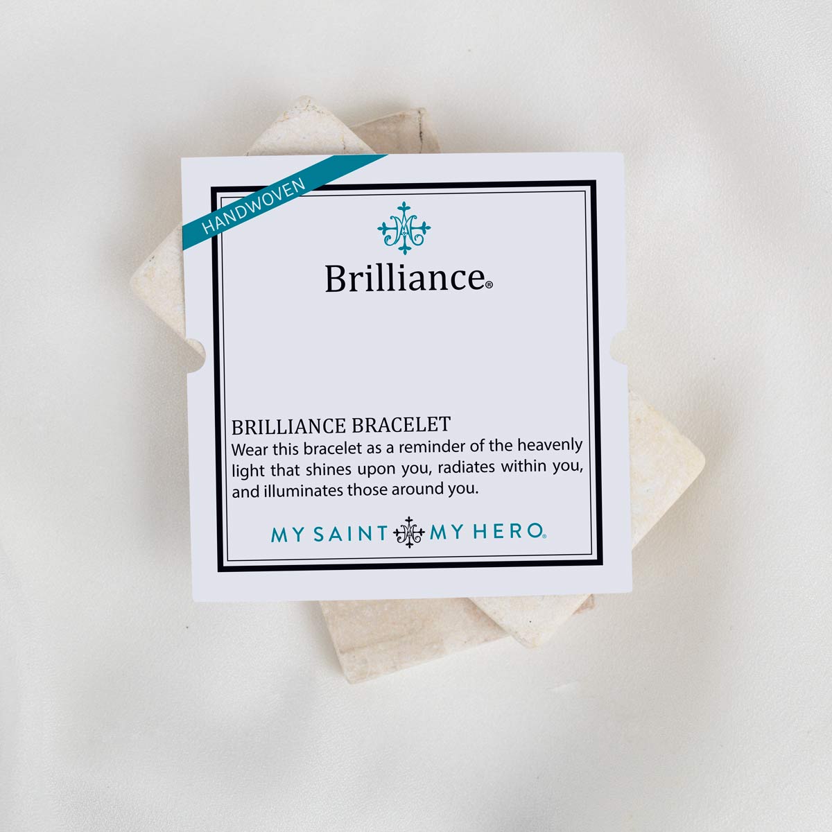 A white square card for the Brilliance bracelet, with a teal 'Handwoven' ribbon and black text, placed on top of several light beige woven fiber squares against a white background.