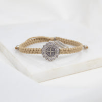 A beige braided cord bracelet with a Benedictine cross medal surrounded by crystals, displayed on a white surface.