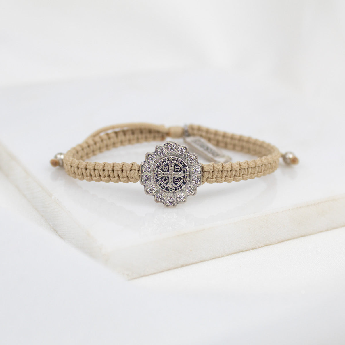 A beige braided cord bracelet with a Benedictine cross medal surrounded by crystals, displayed on a white surface.