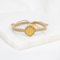 A beige braided cord bracelet with a gold-toned Benedictine cross medal surrounded by crystals, displayed on a white block