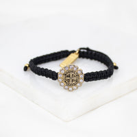 A black macrame bracelet with a central antique gold-toned Benedictine cross medal surrounded by crystals, displayed on a clean white surface.