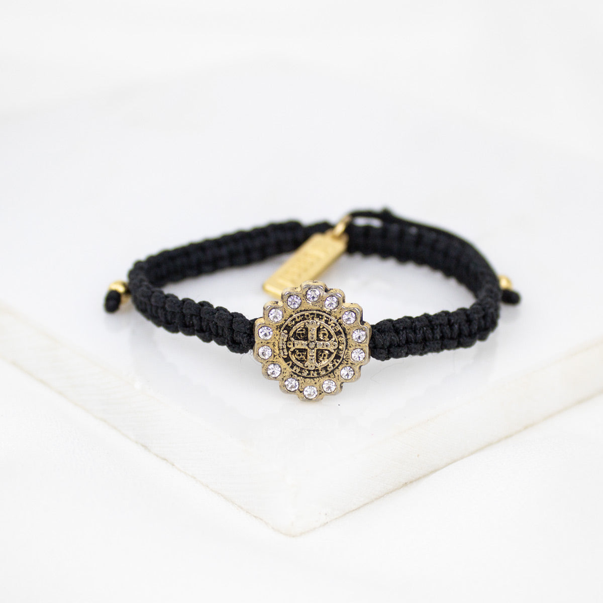 A black macrame bracelet with a central antique gold-toned Benedictine cross medal surrounded by crystals, displayed on a clean white surface.