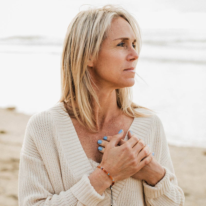 Writer Sara Schulting Kranz wearing Forgive gemstone dainty bracelet with her hands over her heart, looking off to distance, foggy day at beach