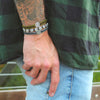 Man with tattoos wearing a stack of two gratitude bracelets and a slate/silver benedictine blessing bracelet