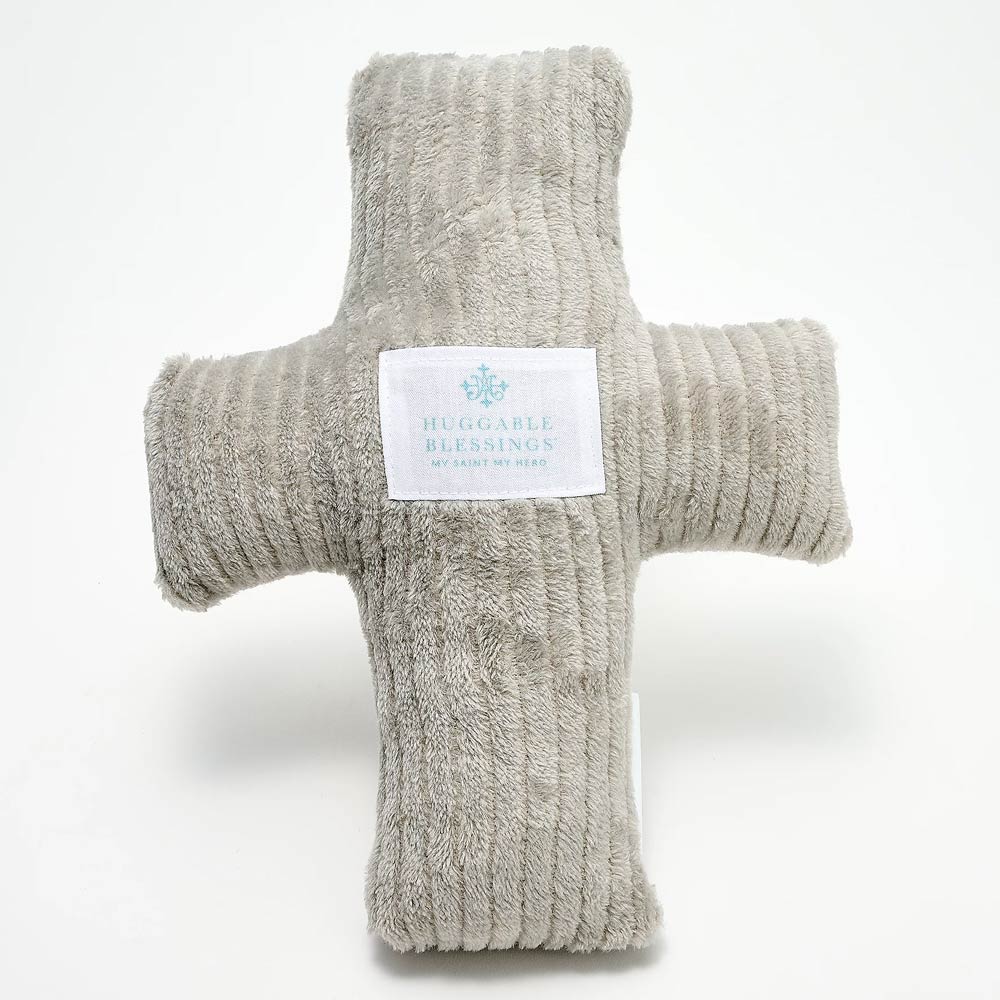A plush, gray weighted pillow in the shape of a cross with a label that reads 'Huggable Blessings'.