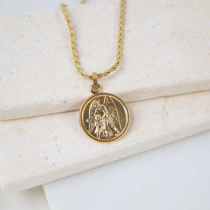 A gold Archangel Raphael Guardian Angel medallion necklace resting on a light-colored, textured stone surface. The round medal features a detailed engraving of the angel with wings spread, holding a staff. The medallion is attached to a matching gold rope-style chain.