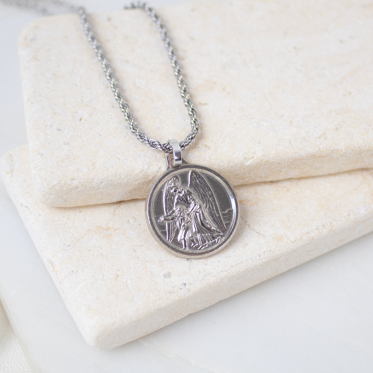 A close-up view of a silver Archangel Raphael Guardian Angel medallion necklace resting on a light-colored, textured stone tile. The round medal features a detailed engraving of the archangel holding a staff, with wings spread wide. It is attached to a matching silver rope-style chain.