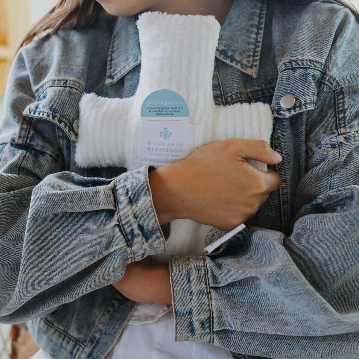 Person holding a white plush cross Huggable Prayer pillow wearing a denim jacket.