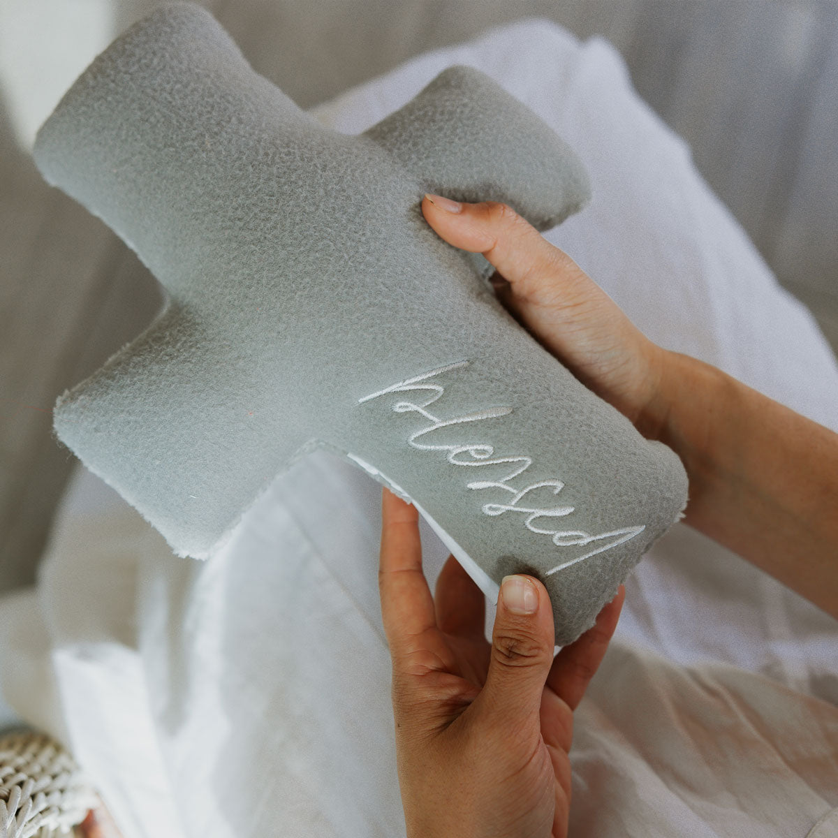 Hand holding a gray cross prayer pillow with 'blessed embroidery against a neutral background