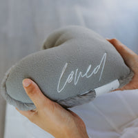 Gray plush pillow with 'loved' embroidery held by a person against a neutral background