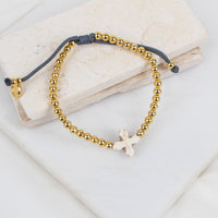 A beaded bracelet with gold-toned round beads, a dark gray adjustable cord, and a small white howlite cross, displayed on stacked light stone blocks.