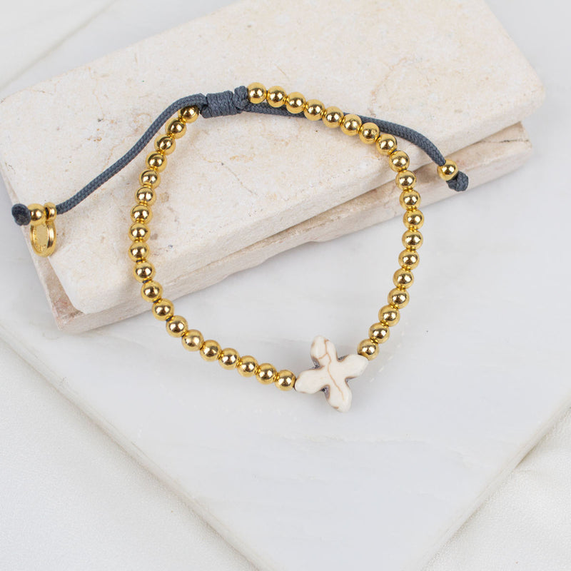 A beaded bracelet with gold-toned round beads, a dark gray adjustable cord, and a small white howlite cross, displayed on stacked light stone blocks.