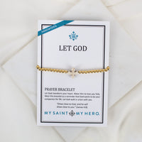 A gold beaded prayer bracelet with a small cross howlite bead, displayed on a white branded card that reads 'LET GOD' and 'MY SAINT MY HERO.' The card is photographed on a soft white fabric background.