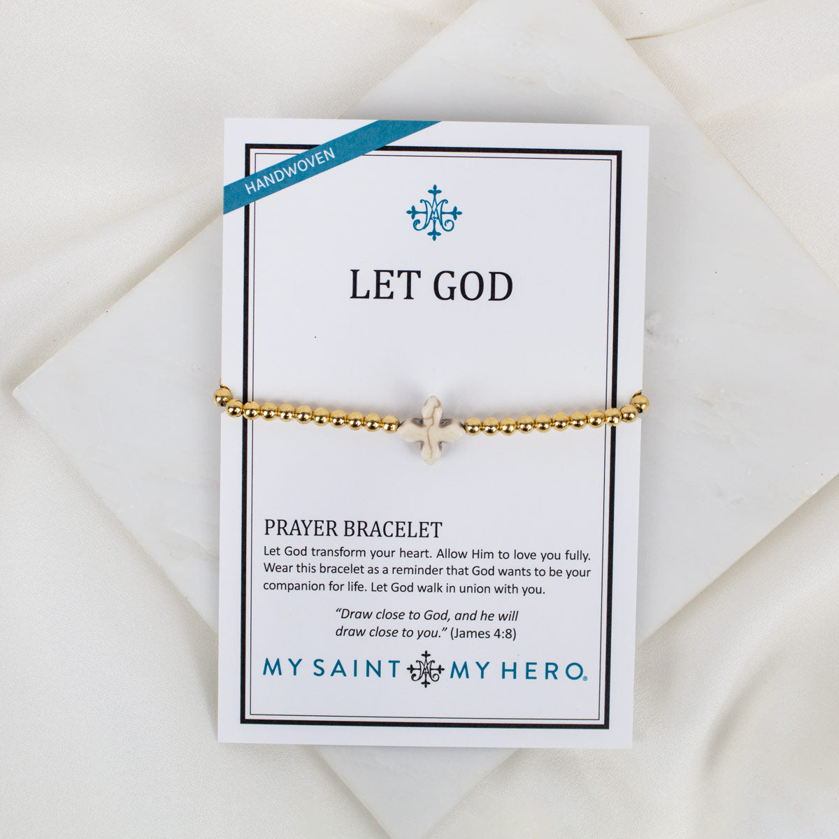 A gold beaded prayer bracelet with a small cross howlite bead, displayed on a white branded card that reads 'LET GOD' and 'MY SAINT MY HERO.' The card is photographed on a soft white fabric background.