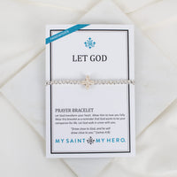 A silver beaded prayer bracelet with a small cross displayed on a white card that reads 'LET GOD' and 'PRAYER BRACELET' with the brand 'MY SAINT MY HERO.' The card is photographed on a soft white fabric background.