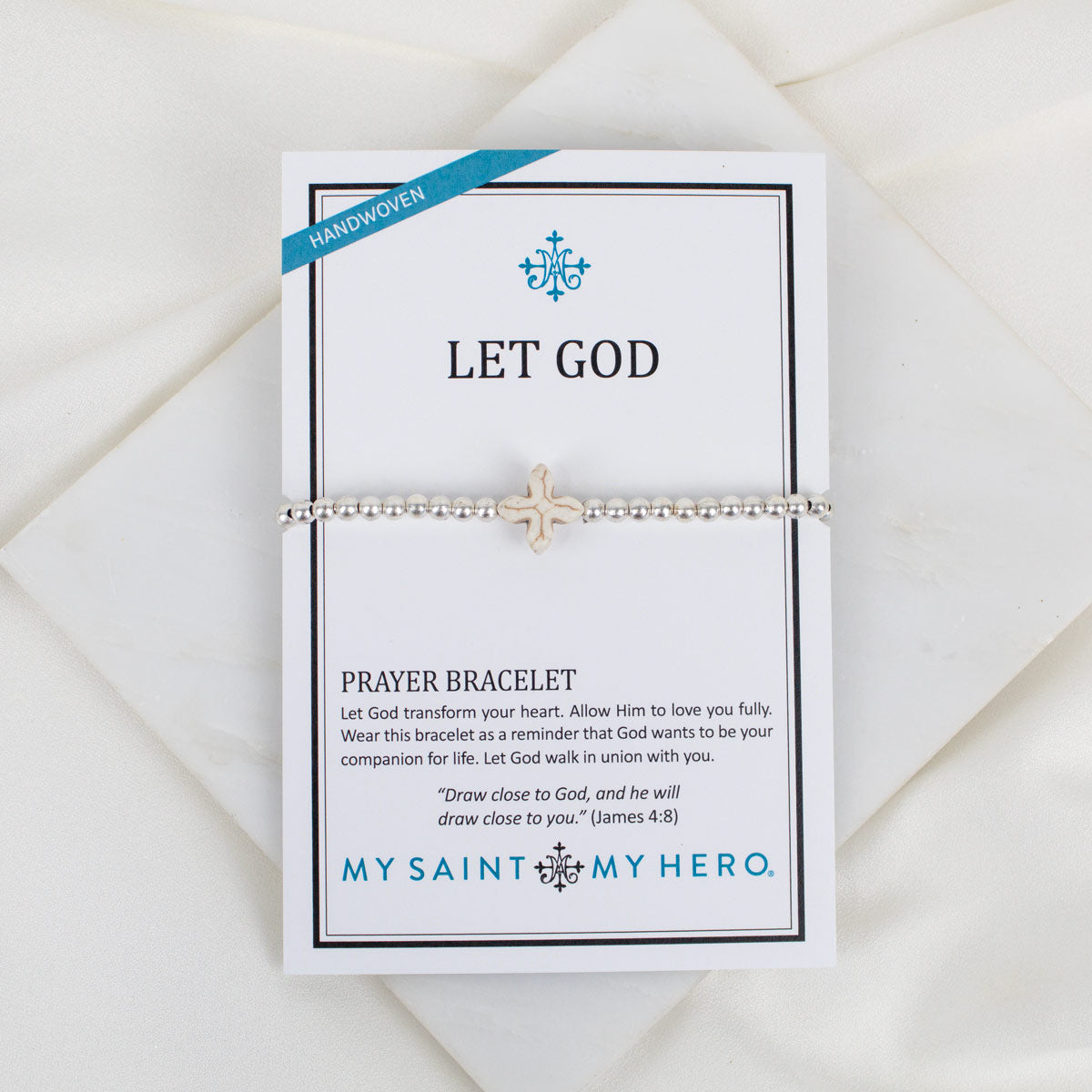 A silver beaded prayer bracelet with a small cross displayed on a white card that reads 'LET GOD' and 'PRAYER BRACELET' with the brand 'MY SAINT MY HERO.' The card is photographed on a soft white fabric background.