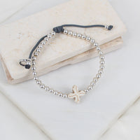 A silver beaded bracelet with a small howlite cross, displayed on a stack of light beige stone tiles.