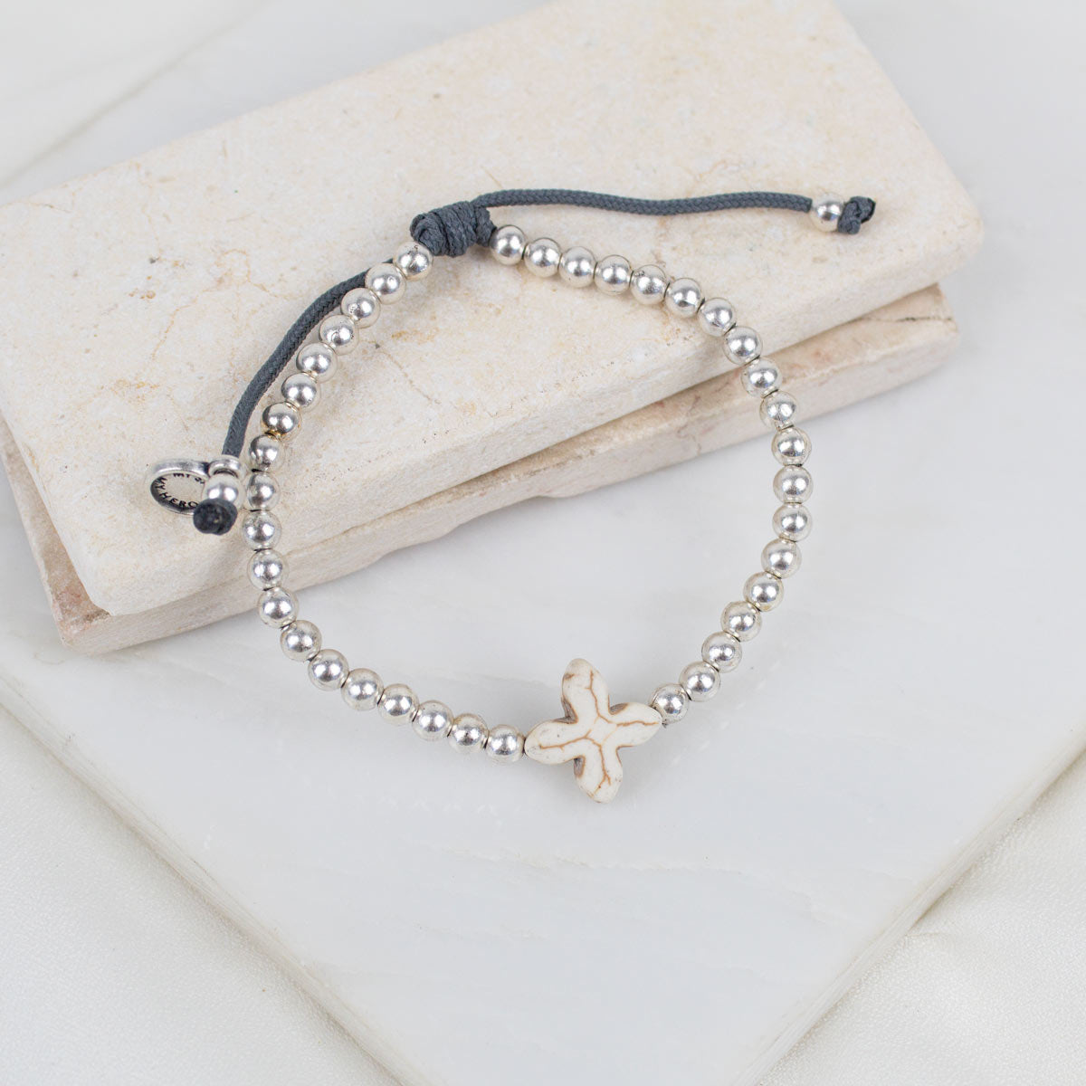 A silver beaded bracelet with a small howlite cross, displayed on a stack of light beige stone tiles.