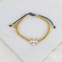 A beaded bracelet with gold-toned round beads, a dark gray adjustable cord, and a small white cross, displayed on a white marble surface.