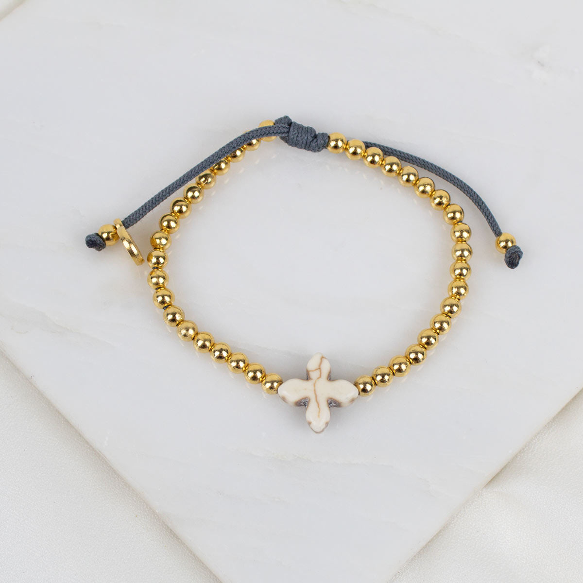 A beaded bracelet with gold-toned round beads, a dark gray adjustable cord, and a small white cross, displayed on a white marble surface.