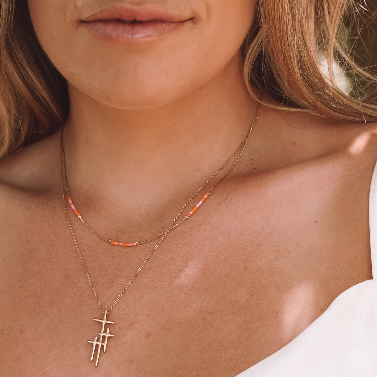 A close-up lifestyle shot of a woman wearing two layered gold necklaces. The shorter necklace features a small beaded accent in orange and white, while the longer necklace features a cross pendant.