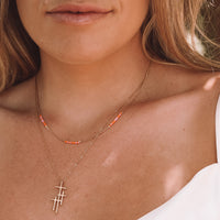 A close-up lifestyle shot of a woman wearing two layered gold necklaces. The shorter necklace features a small beaded accent in orange and white, while the longer necklace features a cross pendant.