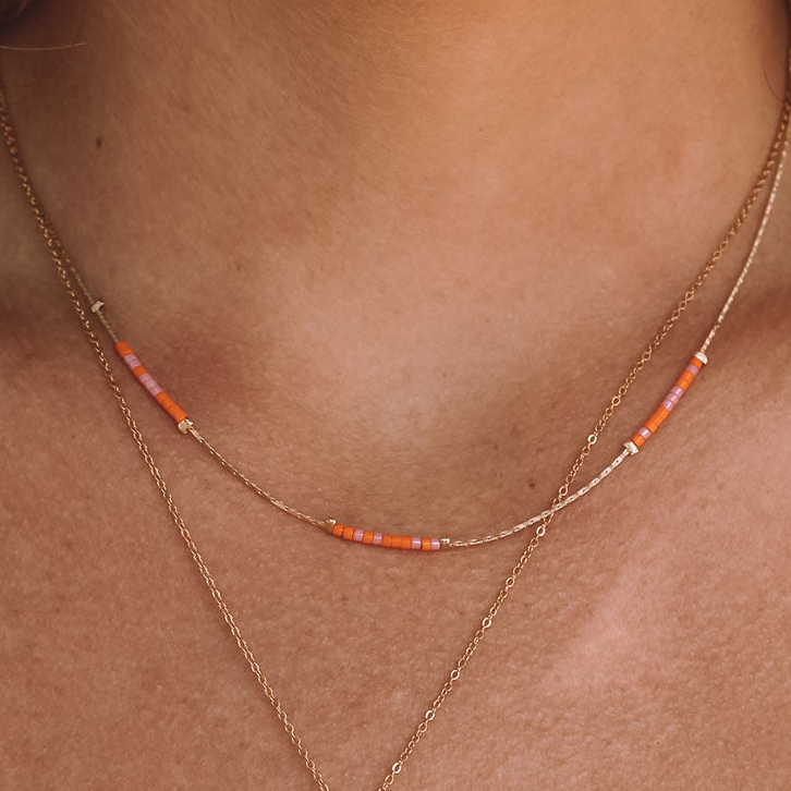 A close-up of a person's neck wearing a delicate layered necklace with small orange and pink beads on a gold chain.