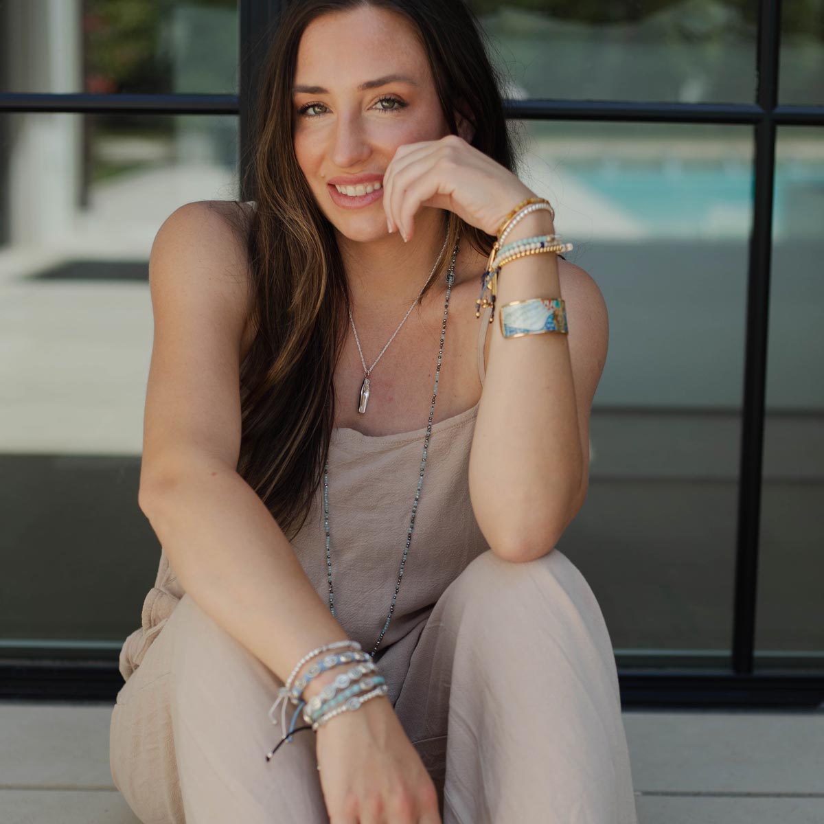 A woman wearing a beige sleeveless jumpsuit and multiple layered bracelets and necklaces, sitting in front of a glass window.