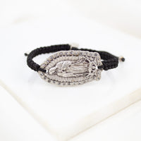 A black braided cord bracelet featuring a silver-toned metal centerpiece with an embossed Our Lady of Guadalupe and small crystals, displayed on a clean white surface.