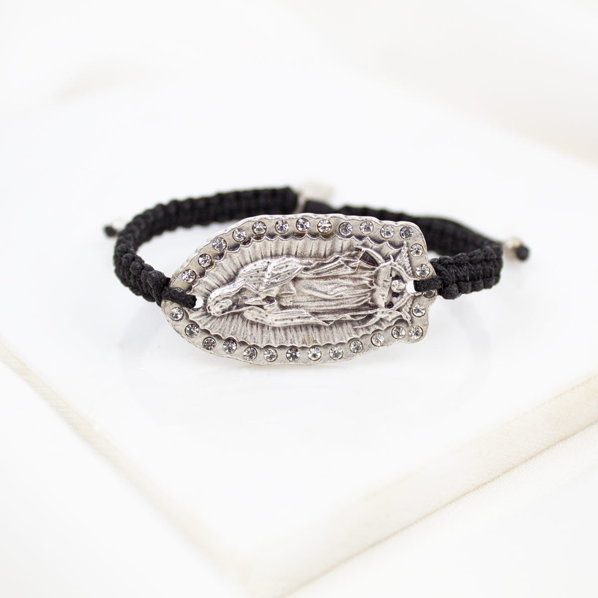 A black braided cord bracelet featuring a silver-toned metal centerpiece with an embossed Our Lady of Guadalupe and small crystals, displayed on a clean white surface.