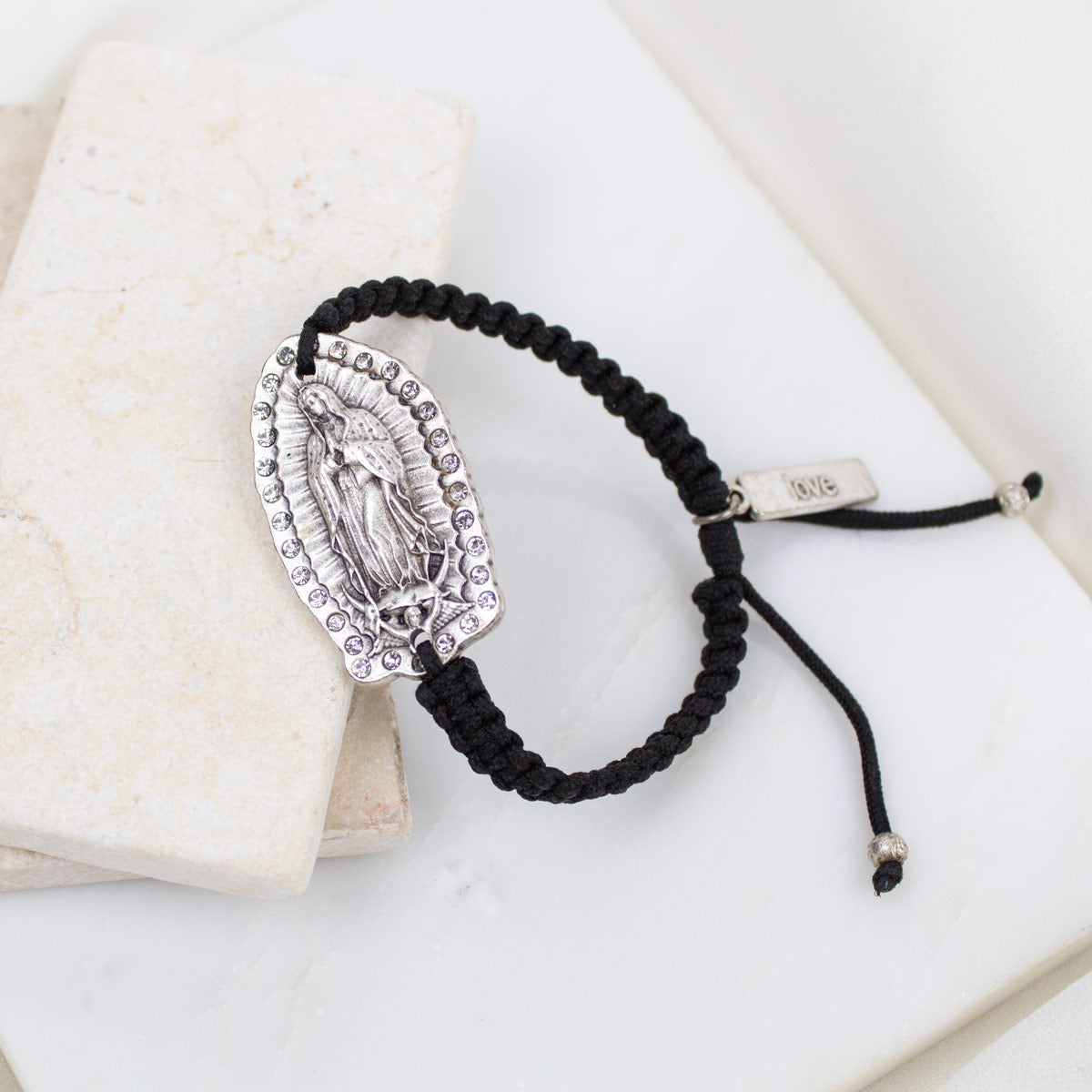 A black braided cord bracelet with a silver-toned Virgin Mary medallion and a small "love" charm tag, styled on a white marble surface next to a cream stone block.