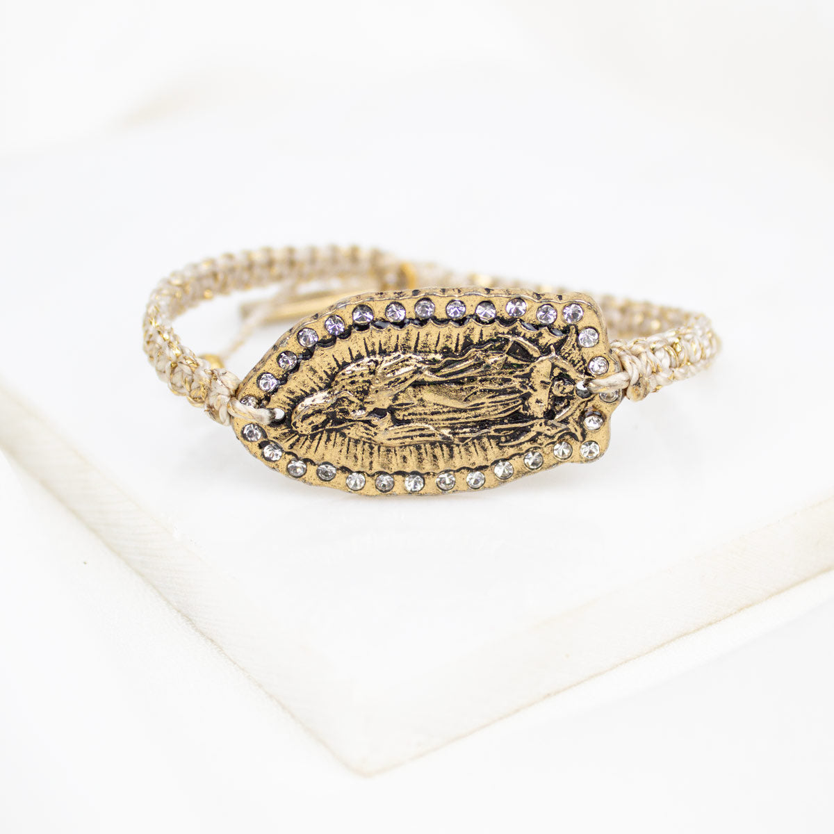 A gold-toned cuff bracelet featuring an embossed religious figure in the center, bordered by small clear rhinestones, displayed on a white textured surface.