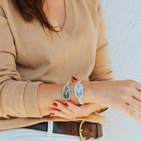 A person wearing a beige sweater has their arms adorned with two bracelets featuring an antique gold or silver-tone Our Lady of Guadalupe medal with crystals.