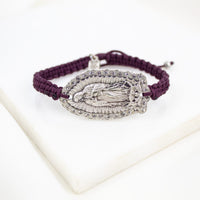 A dark purple woven cord bracelet with a silver-toned Our Lady of Guadalupe pendant, displayed on a white marble block.