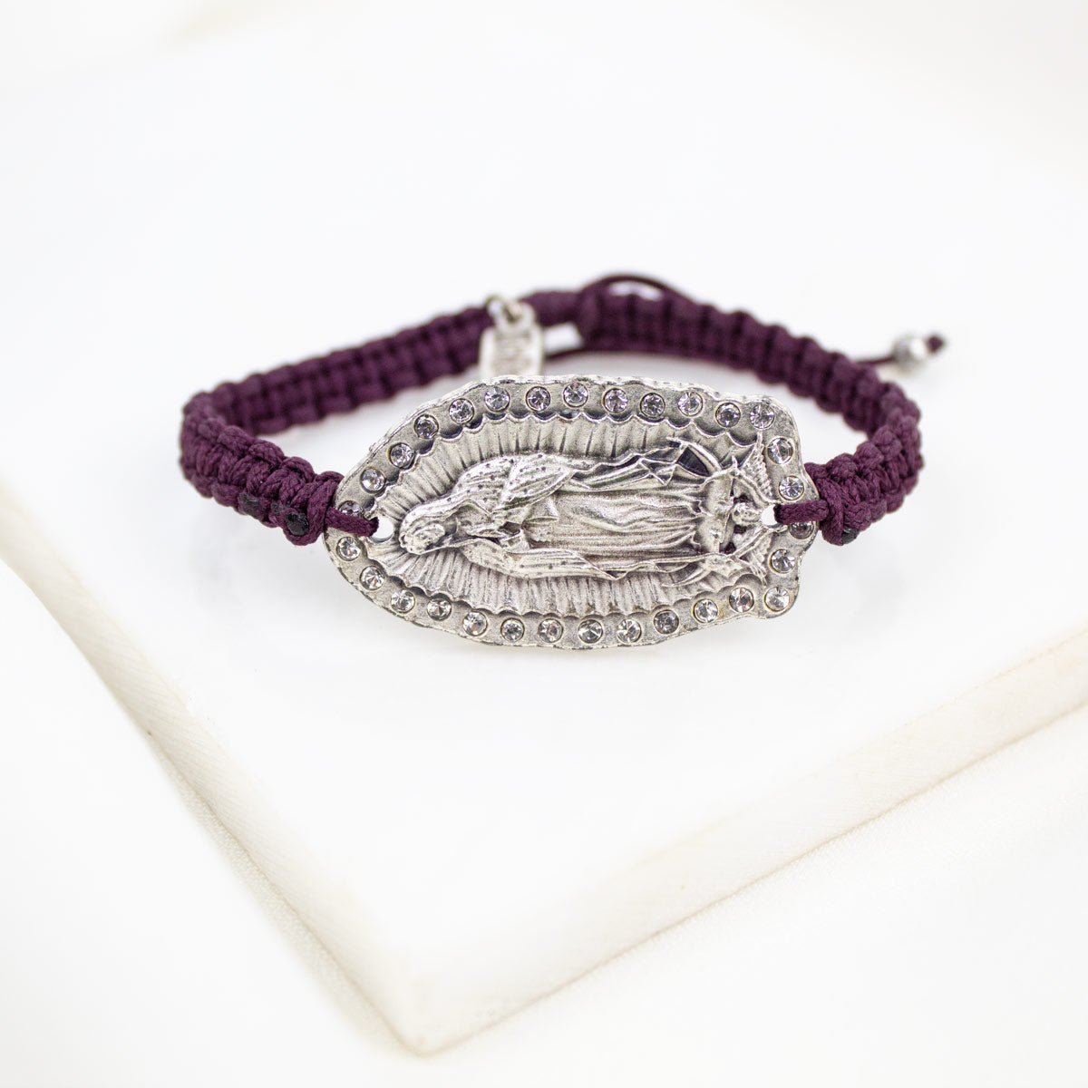 A dark purple woven cord bracelet with a silver-toned Our Lady of Guadalupe pendant, displayed on a white marble block.