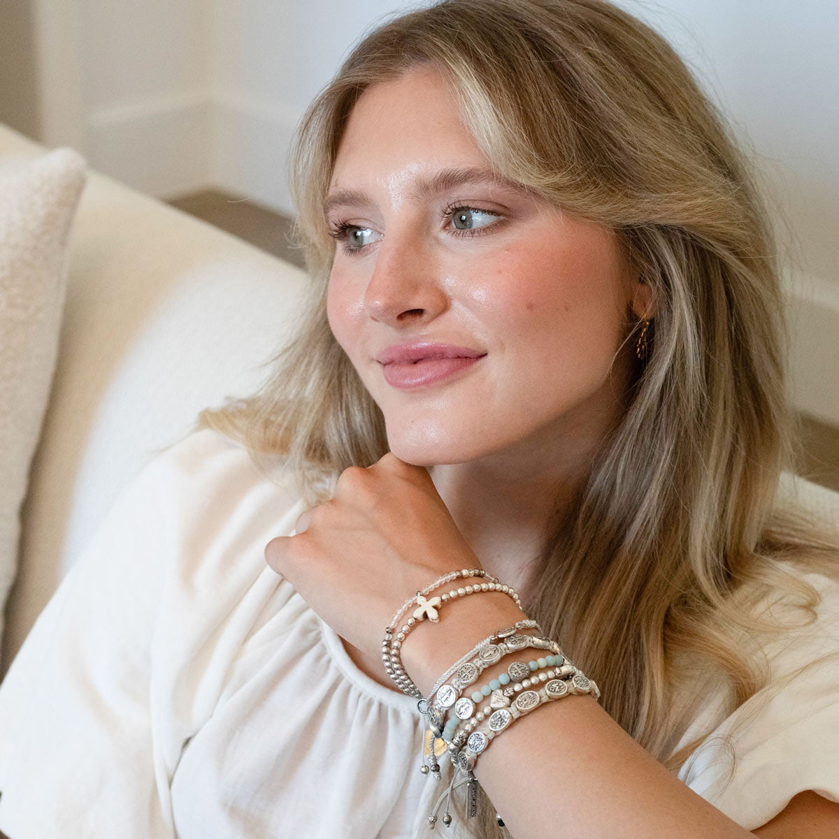 A woman with blonde hair wearing a white blouse and multiple layered bracelets, sitting on a cream-colored sofa in a brightly lit indoor setting.