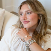 A woman with blonde hair wearing a white blouse and multiple layered bracelets, sitting on a cream-colored sofa in a brightly lit indoor setting.