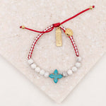 Bracelet with red string, white beads, gold blessed tag, and blue cross charm on a textured beige surface.