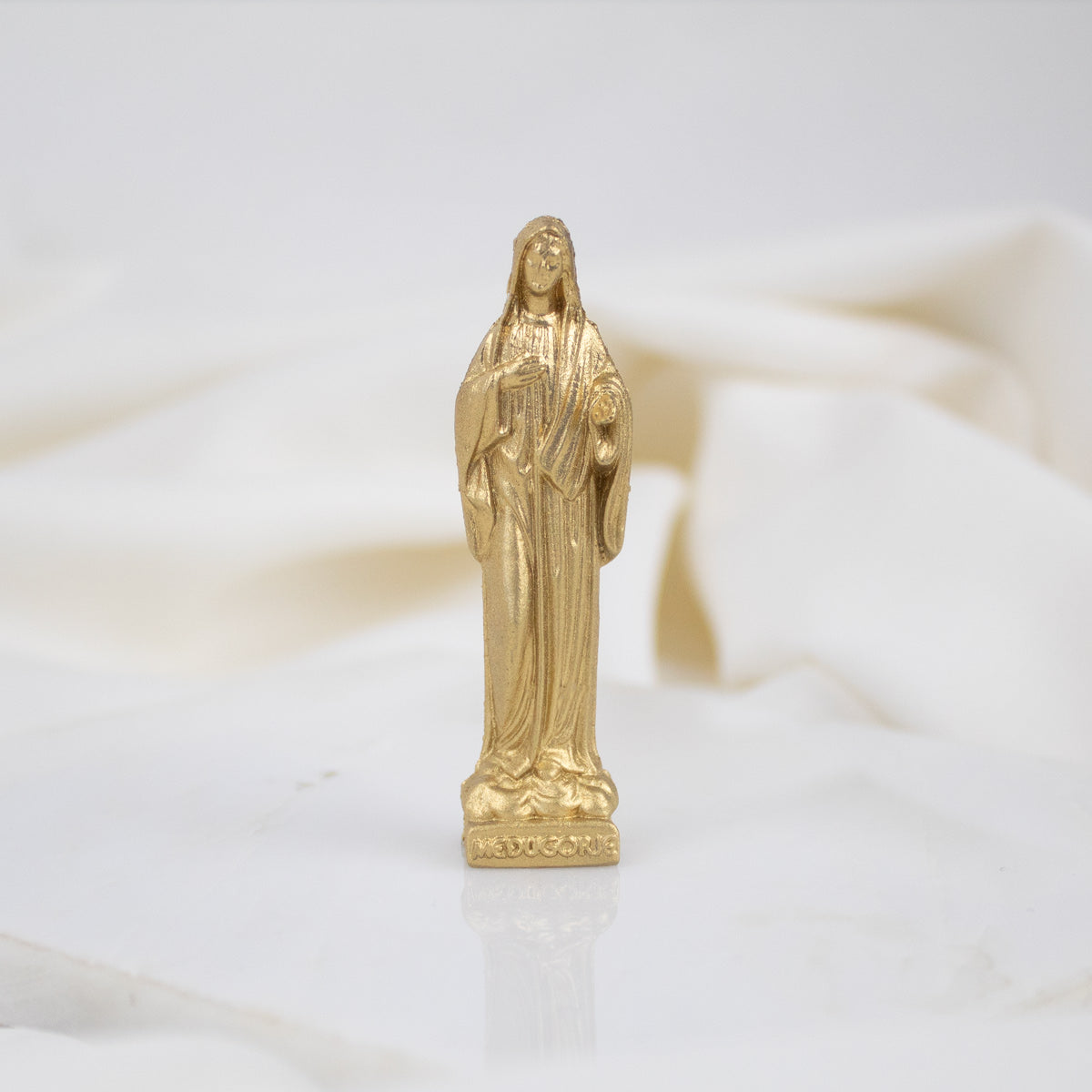 A gold-colored statue of the Virgin Mary with a floral pattern on its base, placed on a white surface.